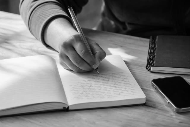 A hand writing in an open notebook on a desk beside a phone